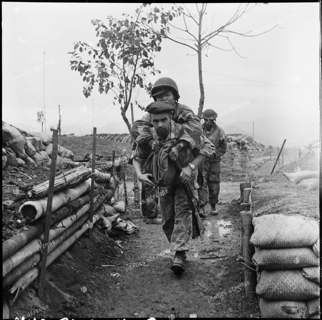 Un parachutiste amène au poste de secours du camp retranché de Diên Biên Phu un camarade vietnamien blessé. En arrière-plan, le poste de commandement du camp retranché.