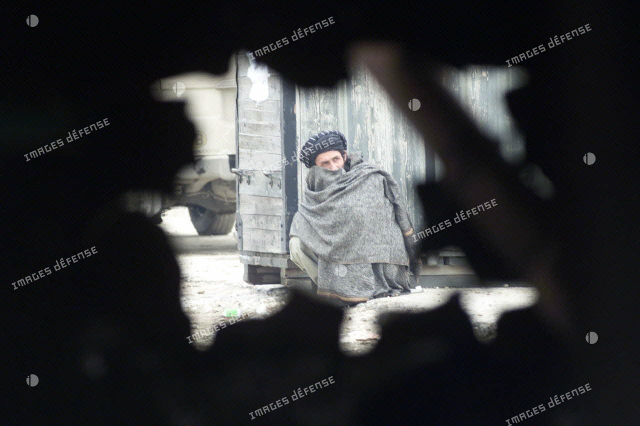 A Mazar e Charif, vue à travers un trou d'éclat d'obus d'un Afghan s'abritant du froid adossé contre une caisse.
