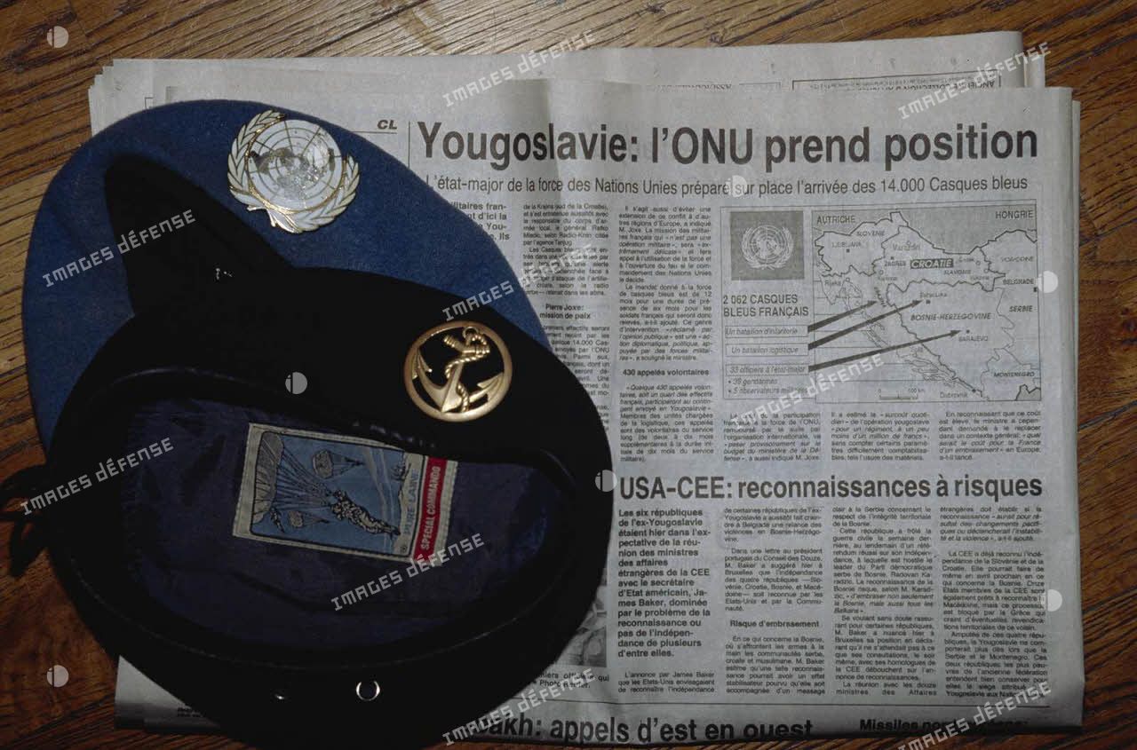 Béret bleu avec insigne ONU et béret Terre avec insigne Troupe de Marine mis en scène sur fond de journal évoquant l'intervention de la FORPRONU en ex-Yougoslavie.