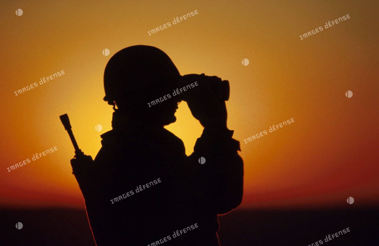 En ZDO (zone de déploiement opérationnel) Olive, un soldat est posté en observation dans le désert, équipé de jumelles, en contre-jour au coucher de soleil.
