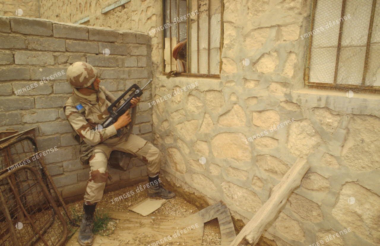 Un groupe de combat du 3e RIMa (régiment d'infanterie de marine) fouille le village d'Al Salman. Un marsouin pointe son fusil d'assaut FAMAS dans la direction d'une fenêtre d'où un habitant sort la tête.