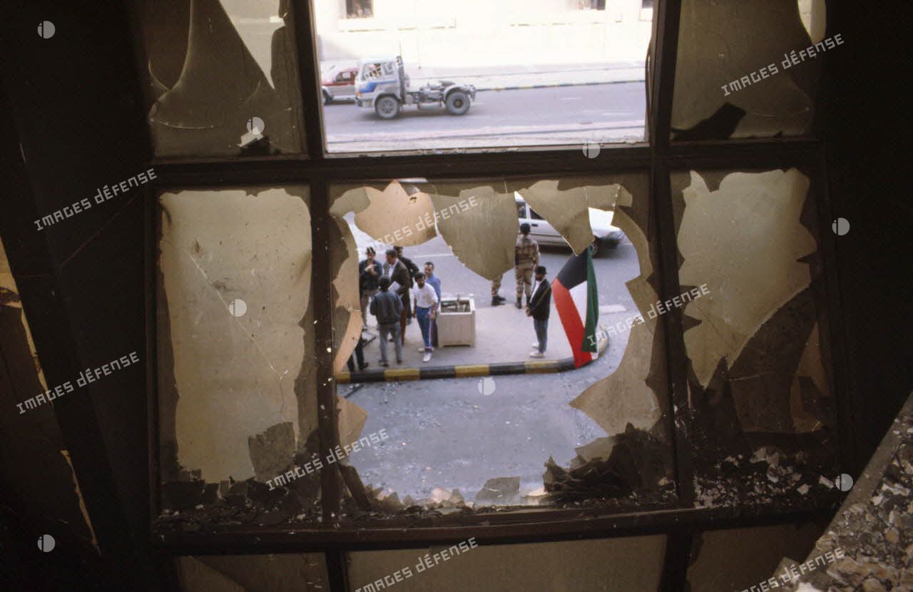 Des civils koweitiens dans la rue vus par une fenêtre cassée depuis l'intérieur carbonisé de l'hôtel Sheraton détruit par les militaires irakiens.