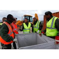 Des militaires du 25e RGA procèdent à l'assemblage d'éléments pour l'édification de gabions sur zone terrassée autour d'un container. En arrière-plan, une tractopelle grecque JCB 3CB utilisée pour le terrassement.