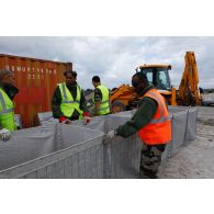 Des militaires du 25e RGA procèdent à l'assemblage d'éléments pour l'édification de gabions sur une zone terrassée autour d'un container. En arrière-plan, une tractopelle grecque JCB 3CB utilisée pour le terrassement.