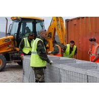 Des militaires du 25e RGA procèdent à l'assemblage d'éléments de structure pour l'édification de gabions sur une zone terrassée autour d'un container. En arrière-plan, une tractopelle grecque JCB 3CB utilisée pour le terrassement.