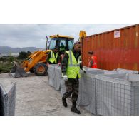 Des militaires du 25e RGA procèdent à l'assemblage d'éléments de structure pour l'édification de gabions sur une zone terrassée autour d'un container. En arrière-plan, une tractopelle grecque JCB 3CB utilisée pour le terrassement.