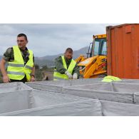Des militaires du 25e RGA procèdent à l'assemblage d'éléments de structure pour l'édification de gabions sur une zone terrassée autour d'un container. En arrière-plan, une tractopelle grecque JCB 3CB est utilisée pour le terrassement.