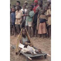 Une mère attend avec son bébé sur un brancard posé devant un groupe de réfugiés lors de l'évacuation sanitaire de blessés de Kibeho par l'élément militaire médical d'intervention rapide (EMMIR). Elle porte une fiche de prise en charge médicale.