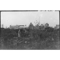 [Soldat posant dans un champ. En fond, village en ruines].<br>