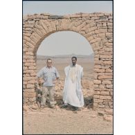Un officier du DMAT d'Atar pose en compagnie d'un guide maure sous l'arche des ruines du décor de cinéma réalisé pour le film "Fort Saganne". [légende d'origine]<br>