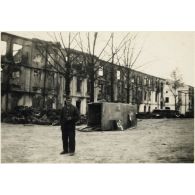 [Un mécanicien allemand pose devant dépôt de réparation. En arrière-plan, des destructions causées par des bombardements. Renversée, une ambulance du service de santé de l'armée de Terre allemande].