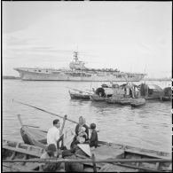Arrivée du porte-avions Arromanches dans le port de Saigon où mouillent des jonques et des sampans.