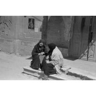 Deux femmes voilées sur le perron d'un bâtiment, à l'arrière-plan une affiche de propagande présentant Adolf Hitler en cyrillique.