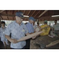 La délégation de membres du gouvernement et d'élus du territoire polynésien visite un atelier d'artisanat tahitien sur l'atoll de Moruroa. Gaston Flosse, président de la Polynésie française (chemisette bleue), et Eugène Bessert, maire de Papara (casquette bleue), examinent des pirogues miniatures et autres objets.