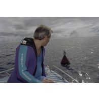 Interview de Jean-Jacques de Peretti, ministre de l'Outre-mer, avant la séance de plongée sous-marine, à bord d'un canot du groupe de prélèvements biologiques du service mixte de surveillance radiologique et biologique (SMSRB) dans le lagon de l'atoll de Moruroa. En arrière-plan, une bouée marque l'emplacement d'un puits de tir d'expérimentation nucléaire sous lagon.