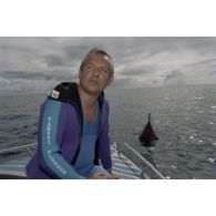 Interview de Jean-Jacques de Peretti, ministre de l'Outre-mer, avant la séance de plongée sous-marine, à bord d'un canot du groupe de prélèvements biologiques du service mixte de surveillance radiologique et biologique (SMSRB) dans le lagon de l'atoll de Moruroa. En arrière-plan, une bouée marque l'emplacement d'un puits de tir d'expérimentation nucléaire sous lagon.