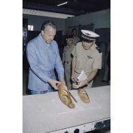 Visite de la boulangerie sur l'atoll de Moruroa. Charles Hernu, ministre de la Défense, goûte une baguette de pain aux côtés du général de corps d’armée aérienne Michel Rouyer, directeur des centres d'expérimentations nucléaires (DIRCEN).