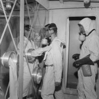 Protégé par un vitrage, un membre de l'équipe de décontamination ôte la tenue de protection d'un personnel à bord du bâtiment de soutien santé (BSS) Rance (A618), spécialisé dans la sécurité radiologique.