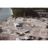 Vue aérienne d'insfrastructures du Centre d'expérimentations du Pacifique (CEP) sur l'atoll de Moruroa : hangars, blockhaus, tour, antenne du poste de commandement de tir (PCT) à Anémone, et un ballon captif jaune dégonflé au sol.