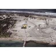 Vue aérienne d'insfrastructures du Centre d'expérimentations du Pacifique (CEP) sur l'atoll de Moruroa : hangars, blockhaus, tour, antenne du poste de commandement de tir (PCT) à Anémone, et un ballon captif jaune dégonflé au sol.