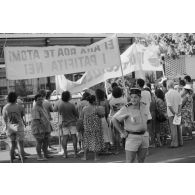 La Gendarmerie nationale à Papeete. Un gendarme devant une manifestation antinucléaire. Banderoles "ei aha roa te atomi i patofita nei" (quel est au juste cet atome qui rayonne ?) et "ia ora te natura" (vive la nature).