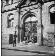 Un soldat français met en joue deux soldats allemands prisonniers avec un fusil d'assaut Sturmgewehr Stg-44, devant la façade d'une école de Fribourg-en-Brisgau.