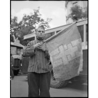 Un déporté politique présente un drapeau à croix de Lorraine sur lequel sont inscrits des noms de camps.