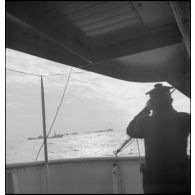 Un marin à bord d'un navire observe un convoi qui progresse sous protection.