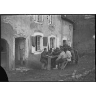 Attablés, des soldats mangent dans la cour d'une ferme.