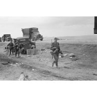 Des fantassins viennent de recevoir leur repas de midi et poursuivent l'avancée parallèlement à des camions Opel Blitz.