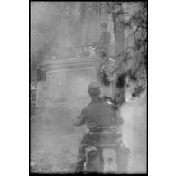 Une colonne de véhicules allemands progresse dans une forêt, au premier plan une moto de la Luftwaffe puis un véhicule sur lequel un insigne d'unité est peint.
