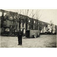 [Un mécanicien allemand pose devant dépôt de réparation. En arrière-plan, des destructions causées par des bombardements. Renversée, une ambulance du service de santé de l'armée de Terre allemande].