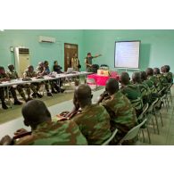 Dans une salle de formation, des stagiaires béninois et sénégalais écoutent le cours sur le droit des conflits armés, dispensé par un commandant ivoirien instructeur, à l'aide d'un rétroprojecteur, en présence d'un journaliste de radio avec son microphone.