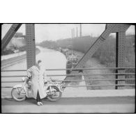 Sur un pont, portrait d'une femme appuyée contre une moto. Au second plan un canal, et une usine en arrière-plan. [légende d'origine]