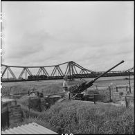 Sur une rive fleuve Rouge, des soldats indochinois sont 	en position de tir sur leur canon antiaérien.