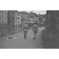 [Années 1930. Deux militaires photographiés de dos marchant au bord d'une rivière dans une ville].