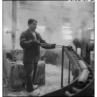 Dans un atelier, un ouvrier manipule des corps d'obus avec une longue pince métallique.