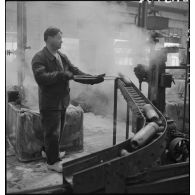 Dans un atelier, un ouvrier manipule des corps d'obus avec une longue pince métallique.