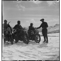 Trois motards à l'arrêt sont photographiés de trois quarts avant, debout un soldat des troupes de montagne discute.
