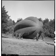 Des aérostiers maintiennent au sol un ballon de protection photographié en plan général de trois quarts avant.
