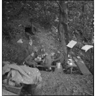 Au bivouac des soldats mangent sur une table de fortune, ils sont photographiés en plan général.