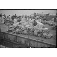 Des sacs de vivres, destinés aux navires d'une escadre, sont stockés sur un quai avant d'être embarqués dans des chaloupes ou vedettes.