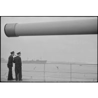Officiers à bord du croiseur léger la Marseillaise observant des mouvements de navires. Au premier plan, un canon de 152 mm d'une des tourelles triples du croiseur.