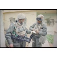 Près d'une maison d'Al Salman, un marsouin du 3e RIMa (régiment d'infanterie de marine) soigne un camarade légèrement blessé à la main, qui fume et porte en collier un émetteur-récepteur TRPP-11. Les deux militaires portent leur équipement individuel : casque F1, brelage, gilet pare-éclats ainsi que leur fusil d'assaut FAMAS et le S-3P (survêtement de protection à port permanent) NBC (risque nucléaire, bactériologique et chimique).