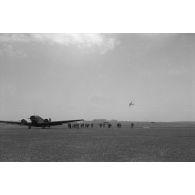 Des troupes s'apprètent à monter dans un avion de transport Junkers Ju-52, en arrière plan dans le ciel de l'aérodrome, un chasseur lourd Messerschmitt Bf-110.