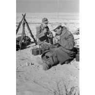 Des soldats mangent près de faisceaux de fusils Kar-98k, de mines antipersonnel, de rouleaux de fils barbelés.
