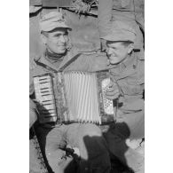 Adossé à un semi-chenillé, un groupe de soldats du DAK autour d'un accordéoniste près d'un camion.