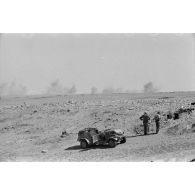 Une voiture Kübelwagen immobile, deux soldats viennent de disposer un poste radio et son antenne, au loin, la poussière provoquée par des explosions.