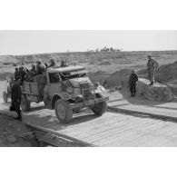 Des véhicules passent un petit pont de bois sous le regard de soldats allemands, ici un camion britannique chargé de fantassins allemands.