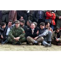 Des responsables Kosovars assis dans l'herbe près de la tribune officielle.